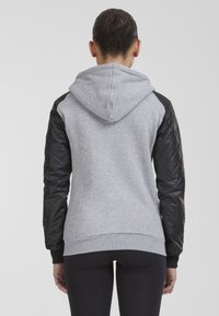 Mujer con el cabello recogido en un moño, vistiendo una sudadera gris con mangas acolchadas negras y leggings negros, de pie mirando hacia atrás contra un fondo plano.