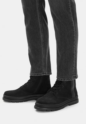 Schwarze Schnürstiefeletten, getragen zu dunkelgrauen Jeans, stehend auf einem hellen, neutralen Hintergrund.