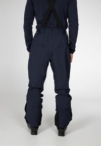 Personne portant un pantalon de neige bleu marine foncé avec des bretelles noires réglables et des chaussures de ski noires, debout devant un fond clair uni.