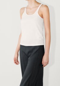 Débardeur blanc moulant à fines bretelles, texture lisse. Porté avec un pantalon gris foncé à coupe ajustée et coutures apparentes.