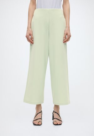 Osoba ubrana w jasnozielone szerokie spodnie o długości do połowy łydki oraz czarne sandały na obcasie z paskami, stojąca na tle jednolitej białej ściany.