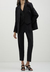 Svart skräddarsydd kostym med en figursydd kavaj, en väst med knappar och avsmalnande byxor. Mjukt tyg med en strukturerad design och minimala detaljer.