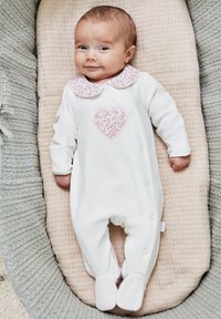 Valkoinen vauvan body, jossa on vaaleanpunainen sydäntulostus, pitsikaulus ja nepparit. Pehmeä materiaali, jalkapohjat, makaa vaaleanbeigeillä pehmustetuilla alustoilla.