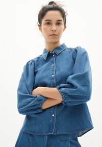 V középkék farmering klasszikus gallérral, elöl gombos záródással és puffos ujjal. Lazán illeszkedő fazon, cuffolt szegélyekkel.