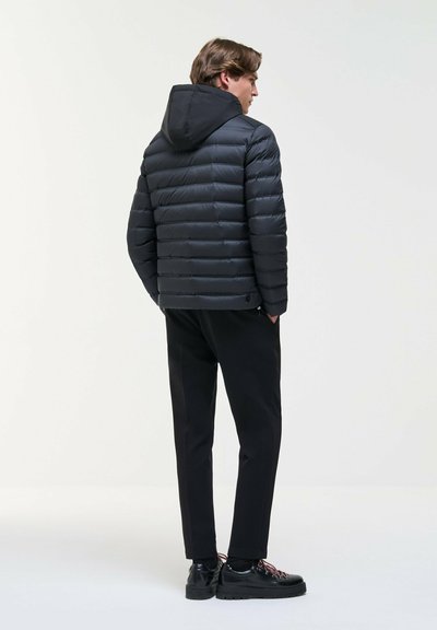 Un homme pose en studio avec une veste noire matelassée à capuche, un pantalon noir et des chaussures à lacets. Il se tient de profil et regarde en arrière.