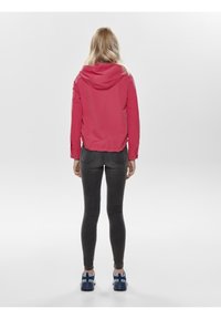Leichte rosa Jacke mit Kapuze, slim fit und geknöpften Ärmelbündchen, kombiniert mit dunklen Hosen und blauen Turnschuhen, von hinten betrachtet.