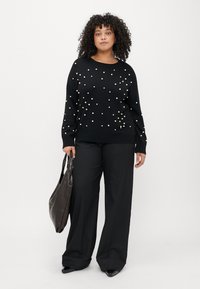 Pulover negru cu accente de perlă dispuse aleator, cu manșete și tiv ondulat, asortat cu pantaloni largi negri și o geantă tote de culoare maro închis.