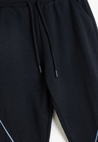 Schwarze Jogginghose mit elastischem Bund, Kordelzug mit schwarzen Enden, seitlichen Taschen und einem hellblauen Akzentstreifen. Weicher, strukturierten Stoff.