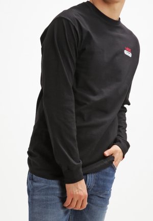 Person iført en sort langærmet skjorte med en lille rød og hvid logo på brystet og blå jeans mod en ensartet hvid baggrund.