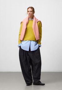 Geel gebreide trui, roze sjaal over de schouders gedrapeerd, lichtblauw shirt, losse zwarte gestructureerde broek, zwarte schoenen, neutrale achtergrond.