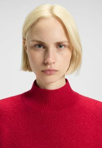 Pull en col roulé rouge avec un design côtelé, fabriqué en maille texturée, doté d'un col montant et de manches raglan.