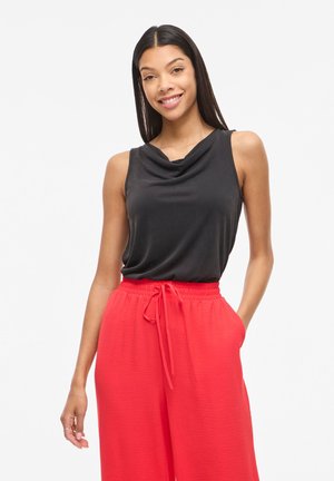 Jonge vrouw glimlachend, draagt een mouwloos zwart topje en losse felrode broek met trekkoord in de taille, handen ontspannen langs haar zij.