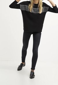 Maglione nero a maglia con una fascia di perline argentate testurizzate sul petto, abbinato a jeans neri slim e scarpe nere con fibbia.
