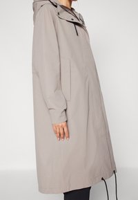 Manteau imperméable gris clair avec capuche, manches longues et coupe droite. Présente des accents noirs et des poches latérales. Cordon de serrage réglable à l'ourlet.