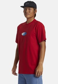 Camiseta de algodón roja con cuello redondo, mangas cortas y un gráfico ovalado azul en el pecho. Llevada con pantalones cortos de denim azul claro.