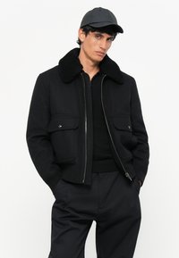 Chaqueta bomber negra con un cuello de piel sintética texturizada, cierre de cremallera y bolsillos de parche en la parte frontal. Lleva puesta sobre un suéter negro y pantalones.