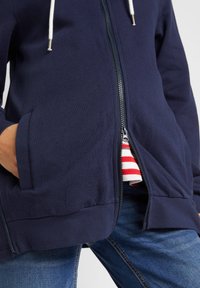 Felpa con zip navy realizzata in tessuto misto cotone, presenta una zip frontale, due tasche laterali e cordini bianchi con estremità metalliche.