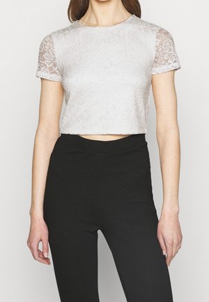 Kvinde iført en hvid blomstret blonde kortærmet crop top og sorte bukser med høj talje, stående mod en ensfarvet baggrund.