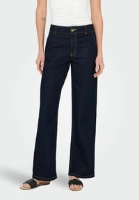 Mörkblå vida jeans med hög midja, prydda med gula sömmar och framknäppning. Matchas med enkla svarta sandaler.