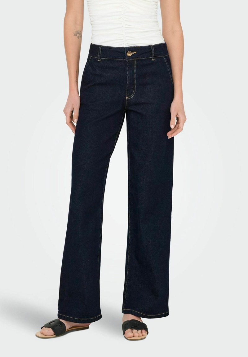 Mörkblå vida jeans med hög midja, prydda med gula sömmar och framknäppning. Matchas med enkla svarta sandaler.