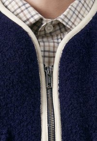 Jūras flīsa jaka ar krēmkrāsas rāvējslēdzēju un apmalēm, pārklāta ar pelēku un brūnu koriģējošo krekli. Tekstūrēta auduma un saskatāma rāvējslēdzēja aparatūra.