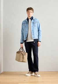 Veste en denim bleu clair sur un pantalon bleu marine foncé, sneakers beige, tenant un sac tan avec des accents marron. Textures lisses et lignes épurées.