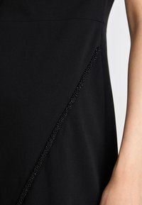 Črna obleka z teksturirano diagonalno črto črnih kroglic ob eni strani, gladka tkanina pa ustvarja eleganten, sodoben dizajn.