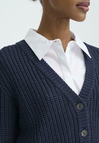 Mörkblå stickad kofta med V-ringning och texturerat mönster, med två knappar. Bärs över en vit skjorta med krage. Mjukt, varmt material.