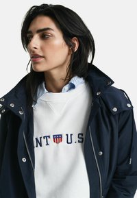 Marinblå jacka med silverdetaljer över en vit sweatshirt med "U.S." i marinblått och en sköldlogotyp, kombinerad med en krage i blåa ränder.