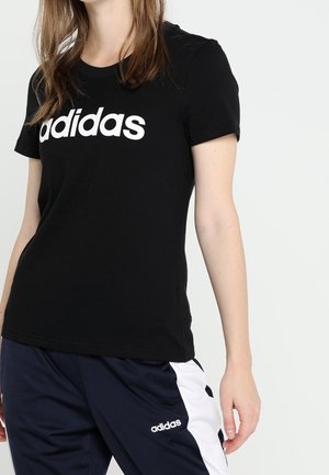 Osoba ubrana w czarną koszulkę Adidas i granatowe spodnie dresowe Adidas z białymi bocznymi paskami, stojąca na jednolitym tle.