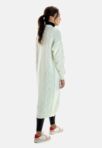 Jersey blanco largo con detalles de punto trenzado, puños acanalados y aberturas laterales, usado sobre leggings negros y zapatillas blancas con acentos rojos.