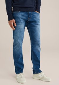 Vyras, vilkintis mėlynus džinsus, tamsiai mėlyną megztinį ir baltus sportinius batelius, stovi su rankomis kišenėse prieš vientisą foną.