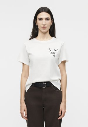 Γυναίκα με μακριά σκούρα μαλλιά, που φοράει λευκό μπλουζάκι με την επιγραφή «le bel été» και μια καρδιά τυπωμένη στο στήθος, συνδυασμένο με σκοτεινό παντελόνι και ζώνη.