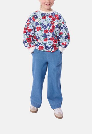 Kind steht mit einem bunten, floral bedruckten Langarmshirt, hellblauen weitem Hose und weißen Sneakers da, mit den Händen in den Taschen.
