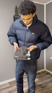Zwarte donsjas met blauwe accenten, grijze beanie met een wit Mickey Mouse-patroon, vastgehouden door een persoon die donkere spijkerbroeken draagt.