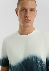 Weißes Baumwoll-T-Shirt mit einem türkisfarbenen Batikmuster am unteren Rand, kurzen Ärmeln und Rundhalsausschnitt.