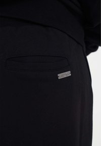 Zwarte joggingbroeken van stof hebben een klein, metalen logoplaatje en een verborgen zak met een platte opening aan de achterkant, waarbij een zachte textuur wordt getoond.