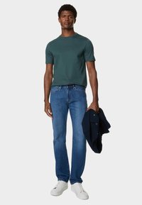 Donkergroen t-shirt met korte mouwen, gecombineerd met blauwe denimjeans. Model houdt een marineblauwe jas vast en draagt witte sneakers. Eenvoudige en casual outfit.