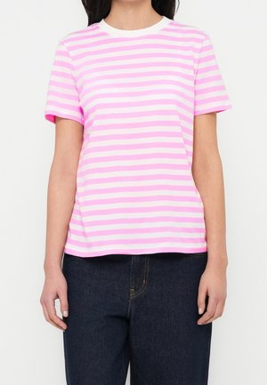 Kvinde iført en lyserød og hvid t-shirt med korte ærmer og horisontale striber, kombineret med mørkeblå jeans, stående foran en ensfarvet baggrund.