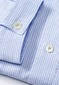 Camicia in gingham a quadri blu chiaro e bianco con due polsini abbottonati e bottoni simili a perle. Tessuto morbido con una texture liscia.