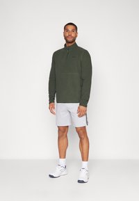 Pullover in pile verde con colletto alto e zip a quarto, abbinato a pantaloni corti grigi chiari con strisce laterali. Il modello indossa scarpe da ginnastica bianche.