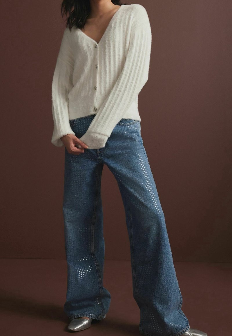 Cardigan blanc à côtes, col en V avec des boutons, associé à un jean large bleu clair avec des motifs perforés et des accents métalliques.