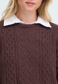 Rjava pletena jopica z strukturiranim vzorcem, okroglo izrezano vratno odprtino in belim ovratnikom, ki štrli izpod nje.