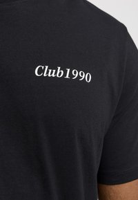 Svart bomull t-skjorte med teksten "Club1990" i hvitt, plassert på venstre brystområde, med myk tekstur og korte ermer.