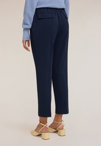 Tmavě modré zúžené kalhoty s jemnou texturou a elastickým pasem, které mají dvě zadní kapsy a zkrácený střih, doplněné o žluté boty na blokovém podpatku.