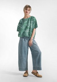 Groene oversized top met bladpatroon en brede mouwen, gecombineerd met lichtblauwe broek met aantrekkoord in de taille en beige instapschoenen.