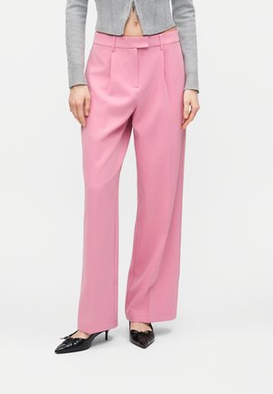 Femme portant un pantalon large taille haute rose, un cardigan gris court, et des chaussures noires à petits talons pointus, debout devant un fond uni.
