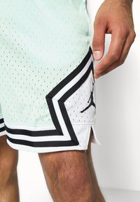 Mátově zelené síťované basketbalové šortky s černými akcenty, bílým logem, třemi černými pruhy na lemu a šikmými bočními rozparky.