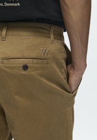 Pantalon en coton marron avec une poche arrière fermée par un bouton noir et un logo "W" brodé. Texture lisse et coupe ajustée.