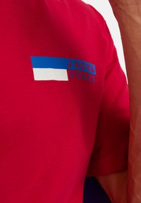 Rotes Baumwoll-T-Shirt mit blauem und weißem Logo, das "JACK & JONES" auf der Brust zeigt. Weiche Textur und kurze Ärmel.